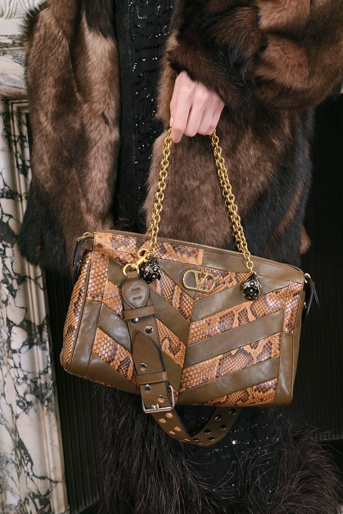 Brown snakeskin‑pattern handbag with chevron leather panels and a gold chain strap, held by a gloved hand over a fur coat edge.