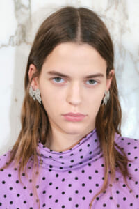 Close-up portrait of a young woman with long brown hair wearing a lavender polka-dot turtleneck and silver leaf earrings.