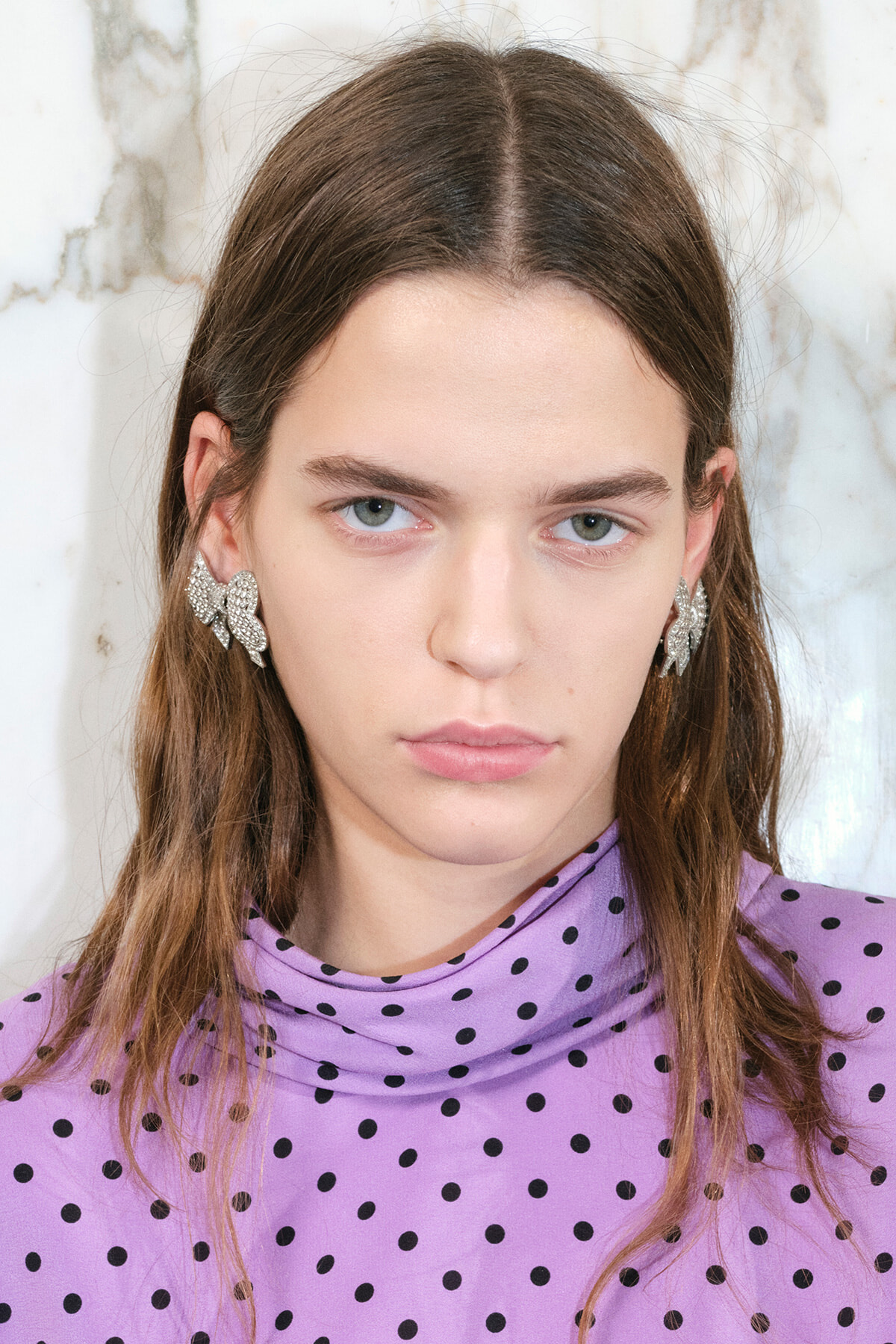 Close-up portrait of a young woman with long brown hair wearing a lavender polka-dot turtleneck and silver leaf earrings.