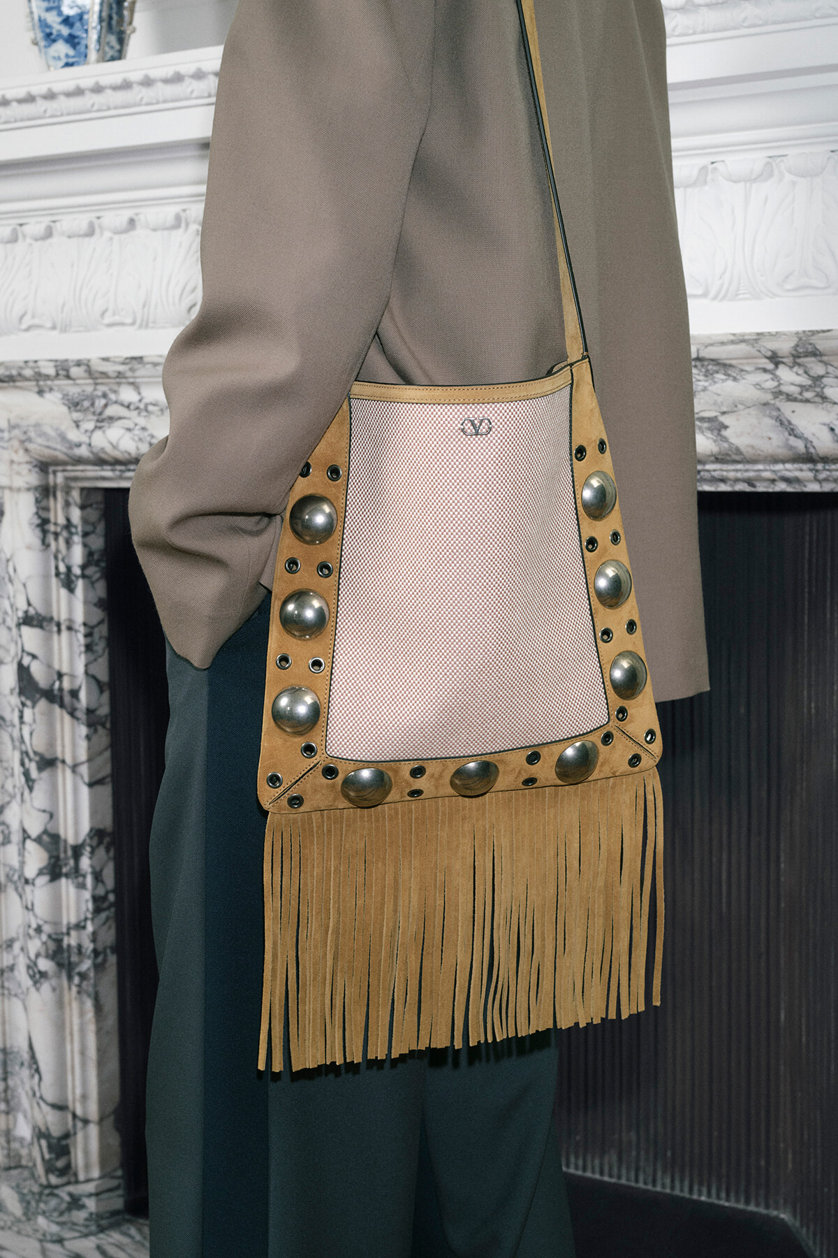 Tan fringe crossbody bag with studded trim worn over a taupe coat in a stylish urban setting.