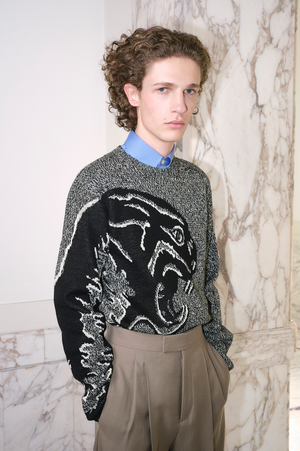 Young man with curly hair in a gray/black patterned sweater over a blue collared shirt, standing against a marble wall.
