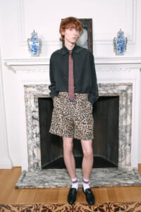 Young male model in an oversized black shirt, red patterned tie, and leopard-print shorts stands in a marble-fireplace setting.