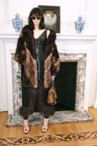 Woman in a dark sequin dress and brown fur coat stands by a marble fireplace, holding a tan handbag and wearing sunglasses.