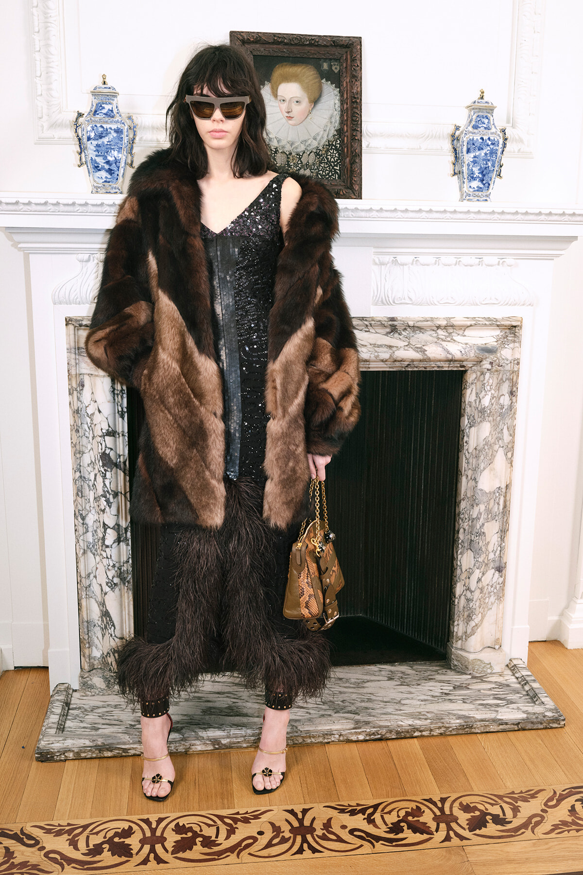 Woman in a dark sequin dress and brown fur coat stands by a marble fireplace, holding a tan handbag and wearing sunglasses.