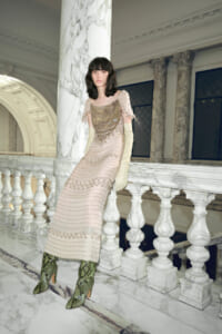 Model in a beige beaded gown with ruffled sleeves and pale gloves leans on a marble railing in an ornate hall awaited by columns and arches.