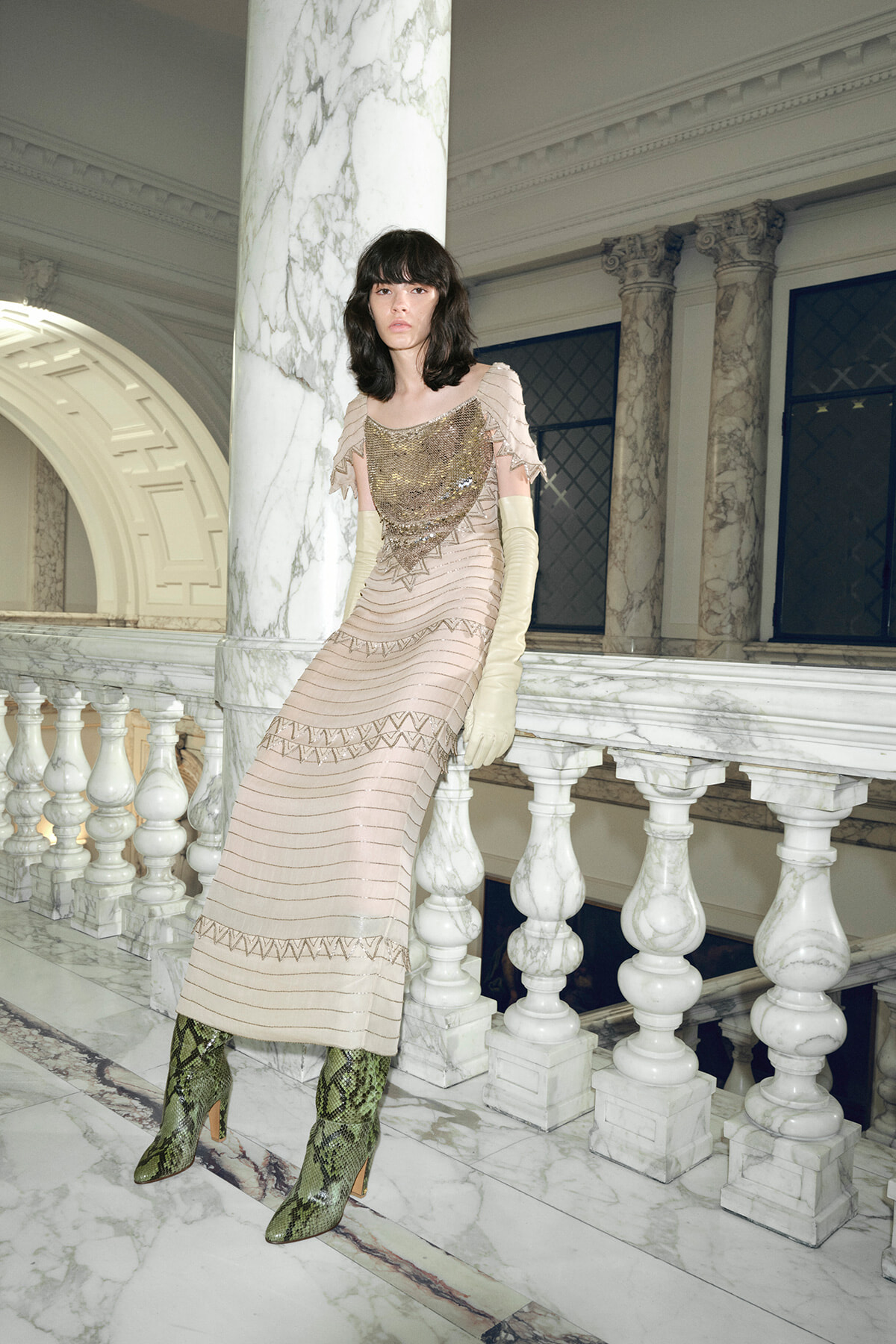 Model in a beige beaded gown with ruffled sleeves and pale gloves leans on a marble railing in an ornate hall awaited by columns and arches.