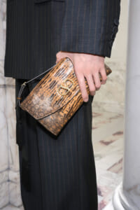 Woman in a pinstriped dark suit holds a brown snakeskin clutch with a gold-tone logo clasp and slim strap.