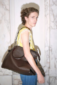 Model wearing yellow lace top and blue jeans carries a large brown leather duffel bag over her shoulder in a marble hallway.