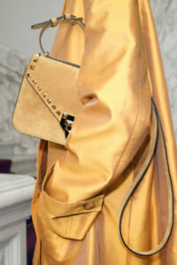 Tan handbag with gold pyramid studs slung over a tan satin trench coat, close-up view of the accessory and strap hardware.