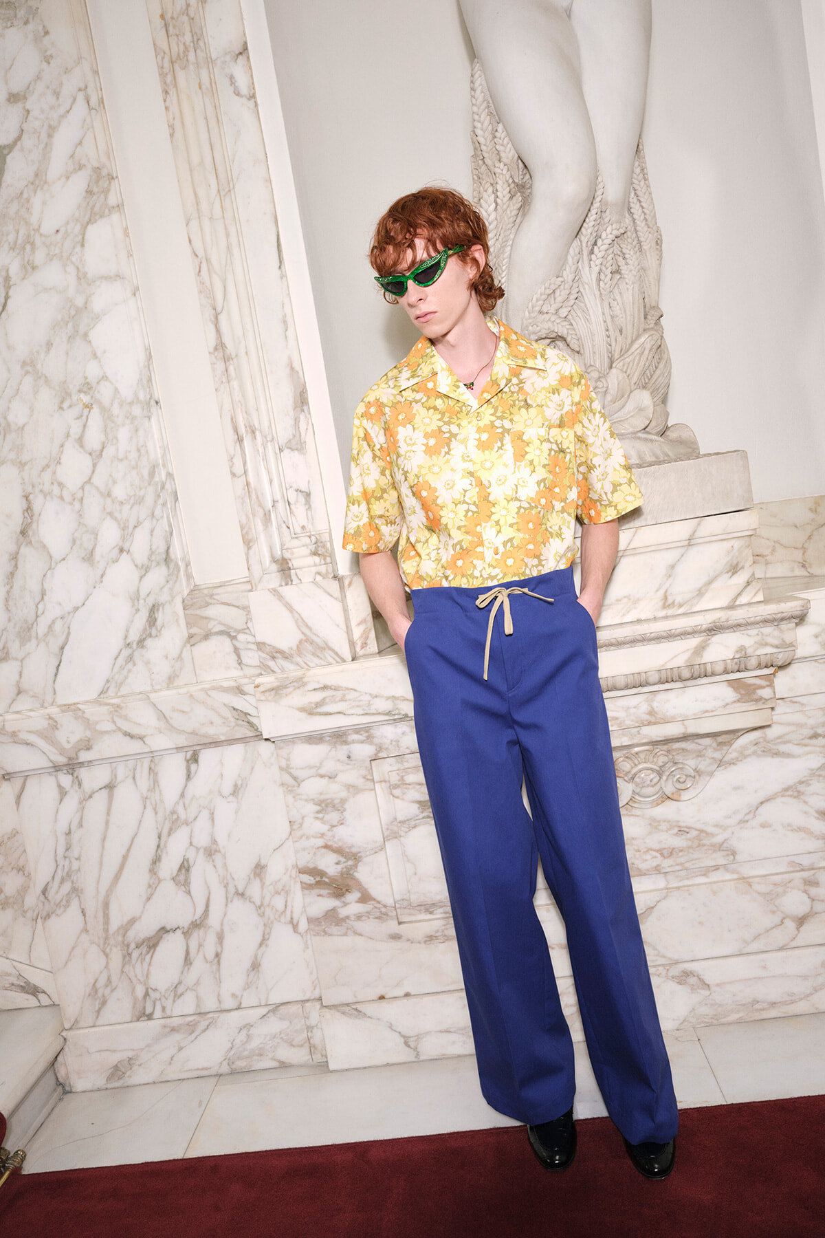 Person wearing a yellow floral shirt and blue drawstring pants, standing against marble steps with green sunglasses.