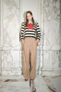Model wearing a black-and-white striped blazer over tan wide-leg pants, red ribbon necklace, standing in marble interior with a brown bag over the shoulder.