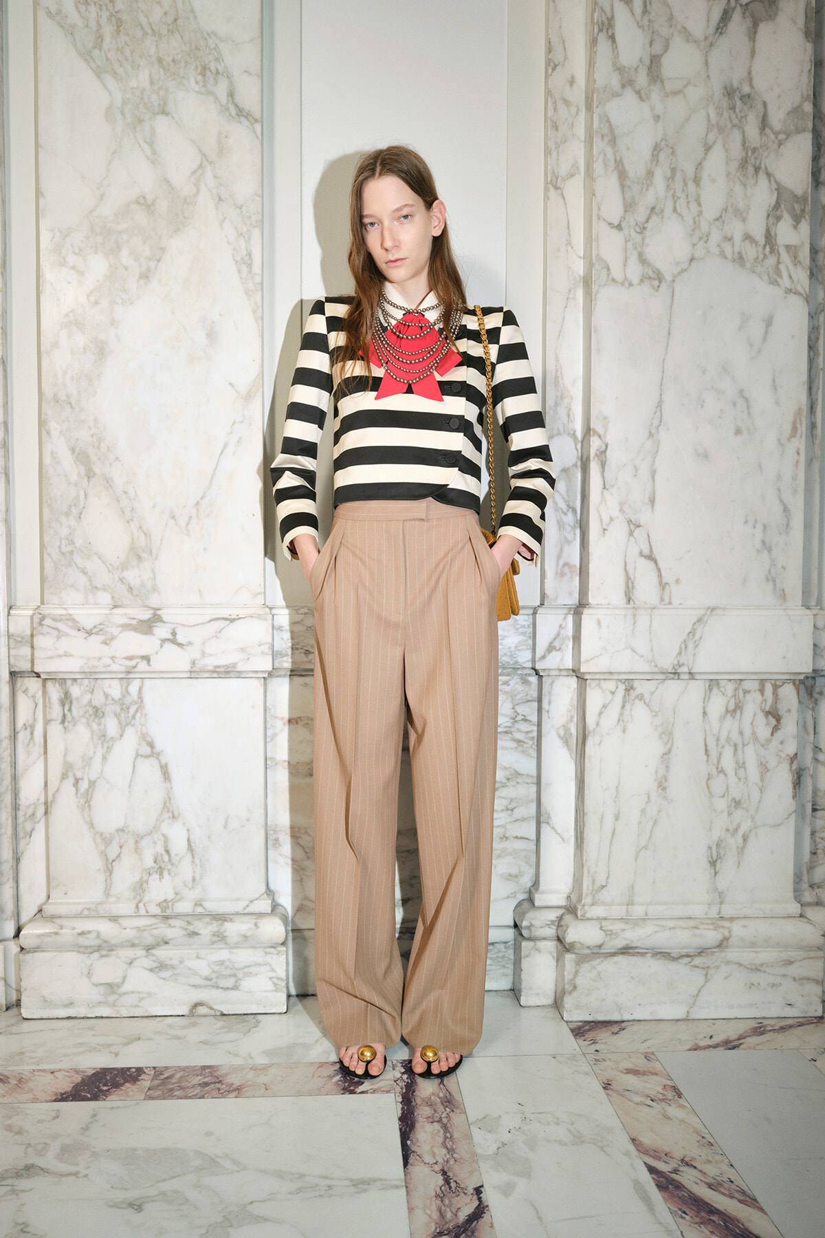 Model wearing a black-and-white striped blazer over tan wide-leg pants, red ribbon necklace, standing in marble interior with a brown bag over the shoulder.