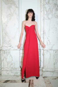 Woman in a strapless red floor-length gown with a knot detail at the bust, standing against marble walls.