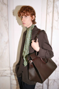 Young person with wavy red hair wearing a brown jacket and green patterned scarf, carrying a brown leather handbag.
