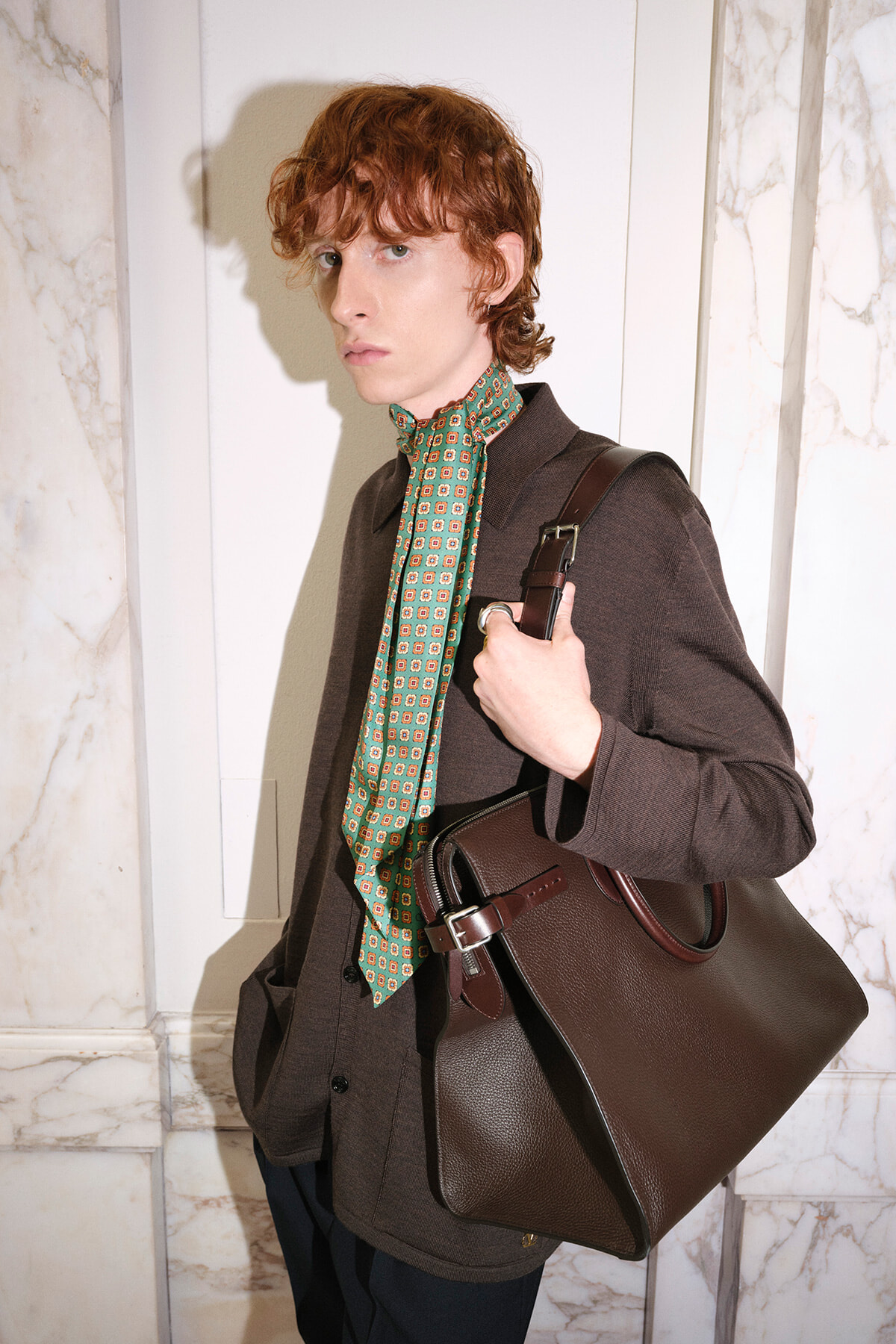Young person with wavy red hair wearing a brown jacket and green patterned scarf, carrying a brown leather handbag.