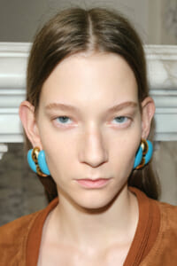 Close-up of a young woman with pale skin, blue eyes, and brown hair wearing bold turquoise and gold hoop earrings.