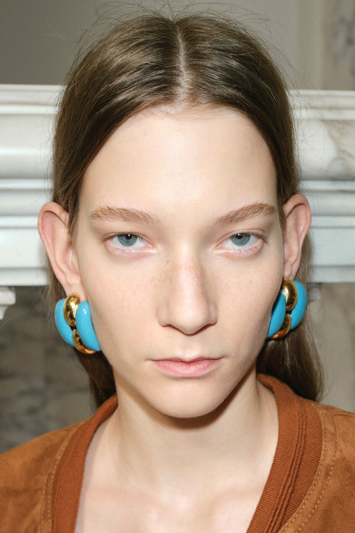 Close-up of a young woman with pale skin, blue eyes, and brown hair wearing bold turquoise and gold hoop earrings.