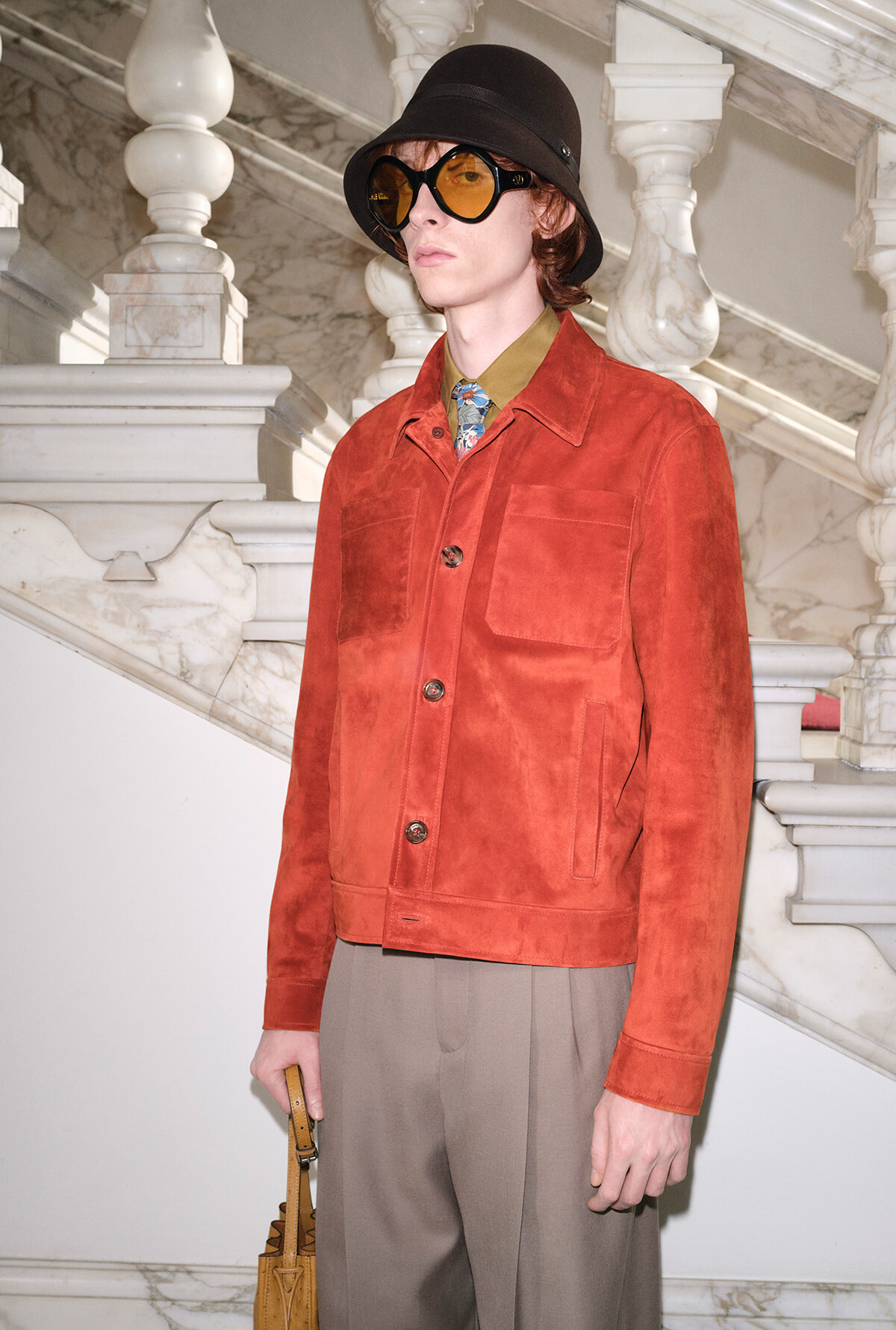 Person wearing a rust suede jacket with large pockets, mustard shirt, patterned tie, black hat and amber sunglasses beside a marble staircase.