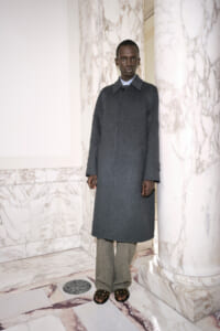 Person standing in a marble-walled hallway wearing a dark gray coat, gray wide-leg trousers, and sandals.