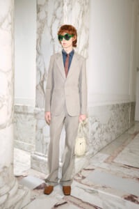 Person in a light gray suit with blue shirt and red tie, wearing large green sunglasses, holding a cream handbag in a marble hallway.