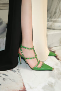 Green strappy high-heeled sandal with gold pyramid studs, worn on a model's foot on a marble floor.