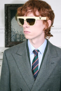 Male model in a gray suit, light blue shirt, striped tie, wearing oversized cream sunglasses indoors.