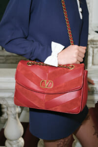 Red chevron-stitched leather shoulder bag with a gold chain strap, carried by a person in a navy outfit with a white bow cuff accessory.