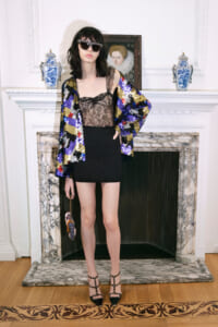 Model posing in a luxurious room, wearing a multicolor sequined jacket over a black lace top and mini skirt, with sunglasses and studded heels by a marble fireplace.