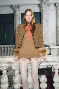 Female model stands on a marble staircase in a leopard-print jacket with fur cuffs, a tan dress, pink bow scarf, and a black crocodile handbag.