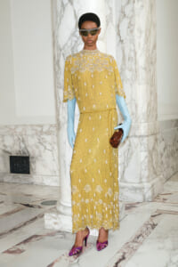 Model wearing a mustard-yellow beaded maxi dress with floral embroidery, sky-blue gloves, dark sunglasses, pink striped heels, and a brown clutch in a marble foyer.