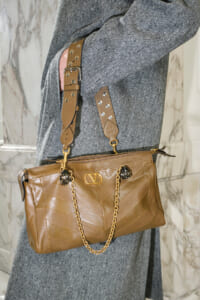 Tan leather handbag with chevron stitching, gold chain strap, and decorative hardware resting against a gray coat.