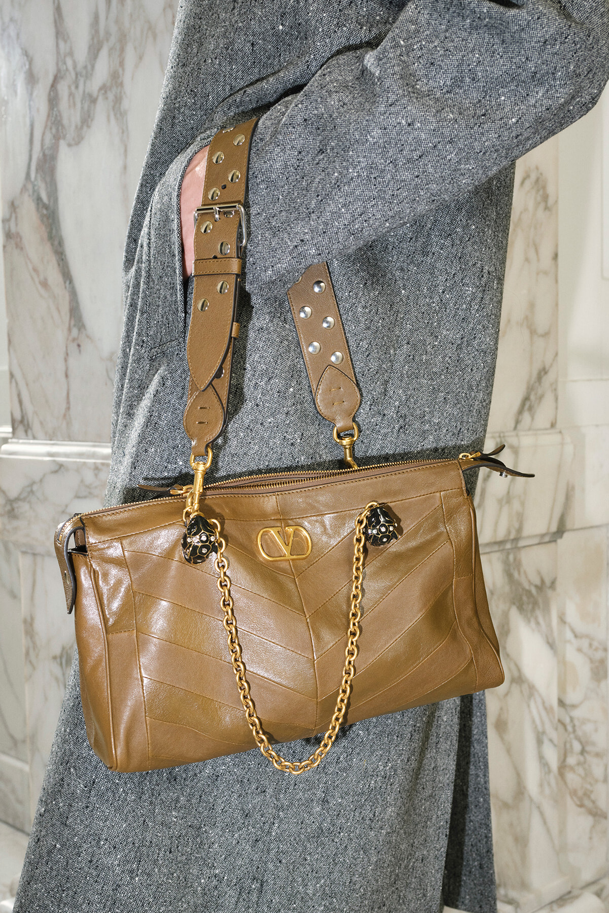 Tan leather handbag with chevron stitching, gold chain strap, and decorative hardware resting against a gray coat.