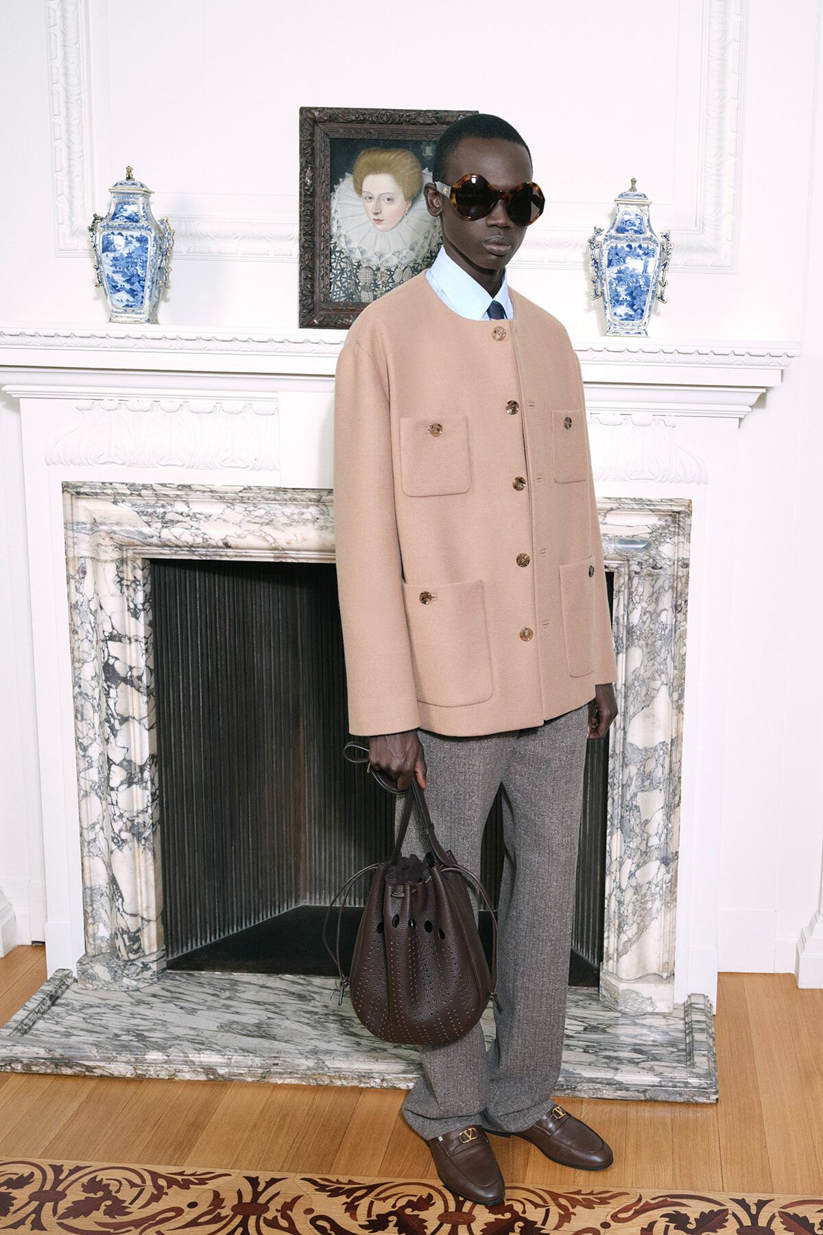 Model wearing a camel coat with large pockets, blue shirt, and oversized sunglasses, holding a brown bucket bag in a luxurious room.