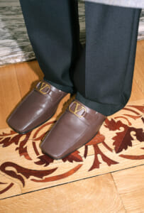 Brown leather loafers with gold-tone buckles, paired with black dress pants, standing on a wooden floor with a decorative rug pattern.