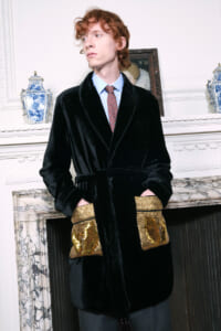 Model wearing a black velvet robe with gold sequin pockets, light blue shirt, red patterned tie, standing beside a marble fireplace in an ornate room.