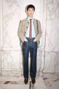 Man in a beige patterned coat over a blue shirt and red tie, wearing high-waisted jeans and brown loafers, standing in a marble hallway with a brown shoulder bag.