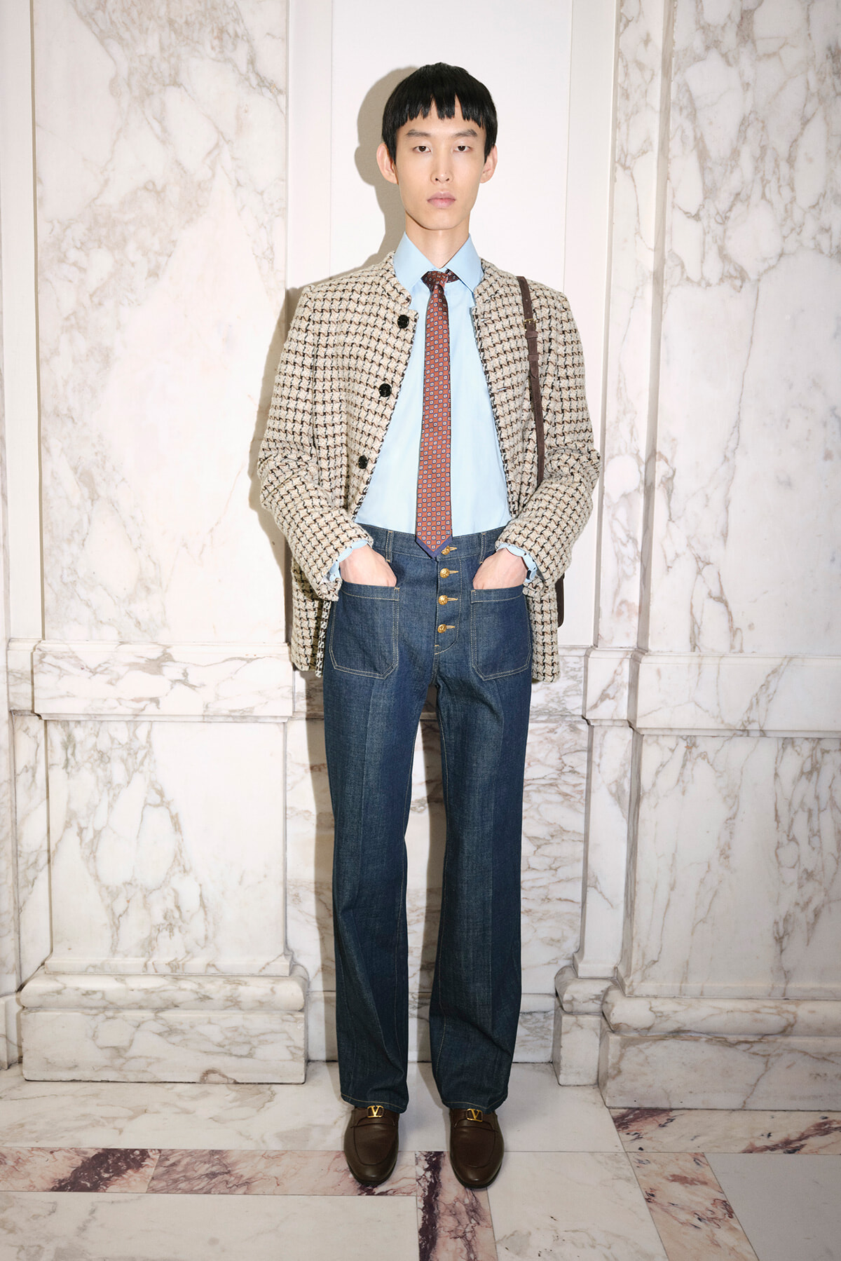 Man in a beige patterned coat over a blue shirt and red tie, wearing high-waisted jeans and brown loafers, standing in a marble hallway with a brown shoulder bag.