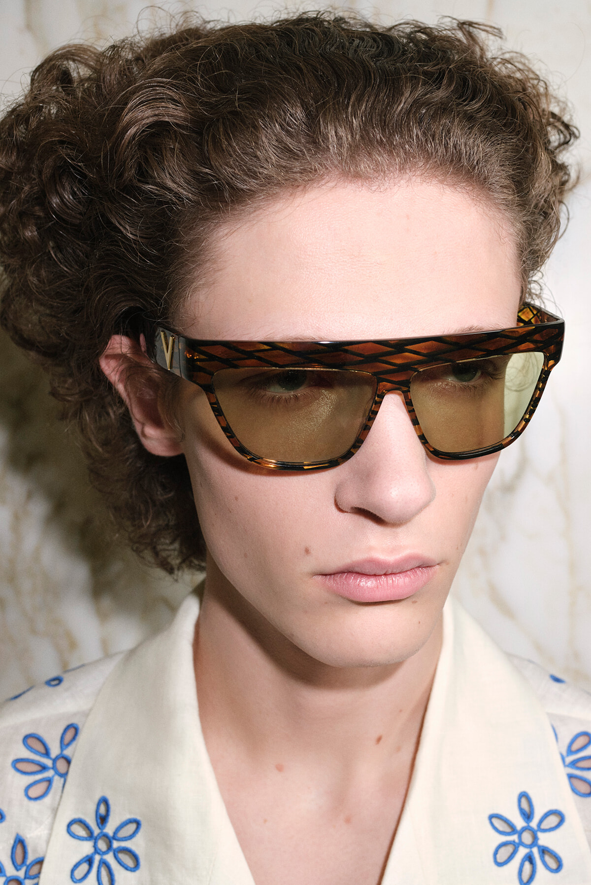 Person with curly hair wearing large tortoiseshell sunglasses and a white shirt with blue embroidery, looking to the side.