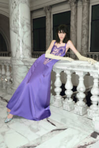 Model in a lavender lace evening gown leaning over a marble railing in a grand marble hall, wearing pale yellow opera gloves.