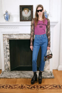 Woman posing indoors by a marble fireplace in a pink 'PANTHER 60' top with lace sleeves, high-waisted jeans, black platform boots, sunglasses, and a floral clutch.