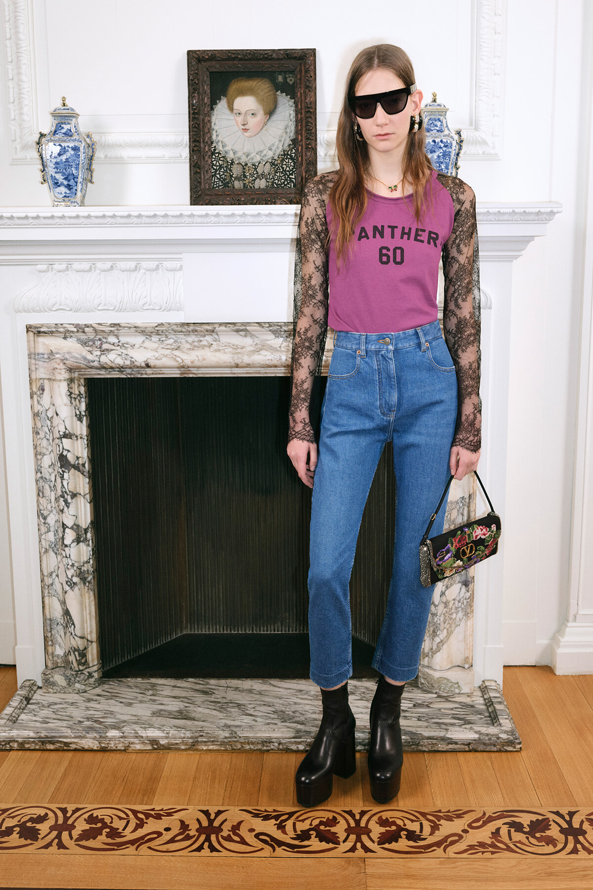 Woman posing indoors by a marble fireplace in a pink 'PANTHER 60' top with lace sleeves, high-waisted jeans, black platform boots, sunglasses, and a floral clutch.
