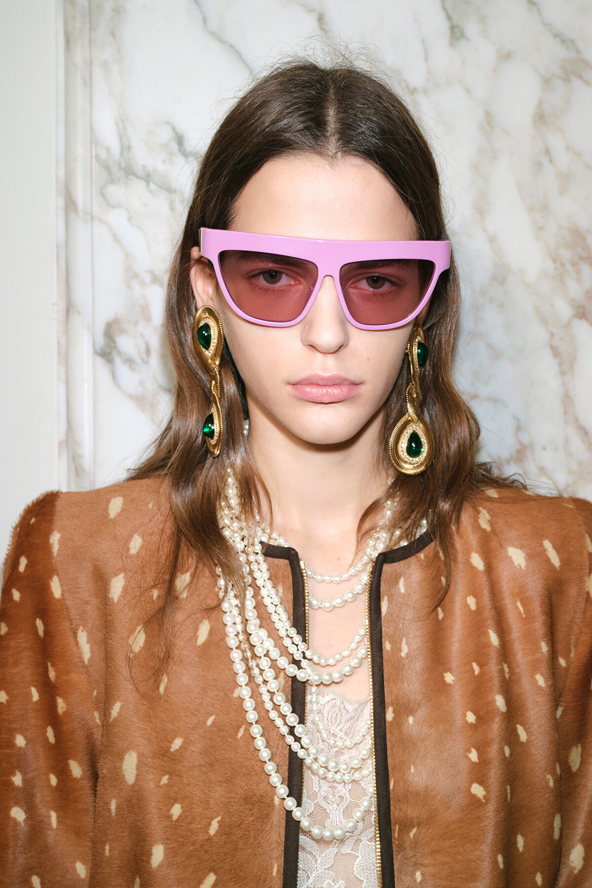 Person wearing oversized pink sunglasses, emerald-green earrings, and layered pearl necklaces over a brown spotted coat.