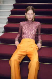 Woman seated on maroon stairs wearing a purple lace turtleneck and bright orange wide-leg pants, with large green and gold earrings.