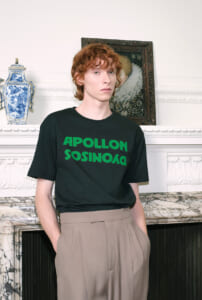 Young man with red hair wearing a black T-shirt with green text, standing in front of a marble fireplace and decorative vase on the mantle.