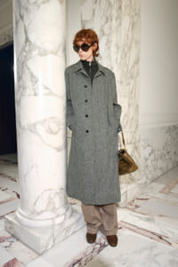 Model in a gray knee-length wool coat, black turtleneck, tan wide-leg pants, brown loafers, and oversized sunglasses beside marble columns in a lobby setting.