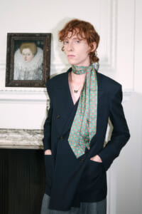 Young person with curly red hair in a navy blazer and teal patterned scarf, standing by a marble mantel with a framed portrait nearby.