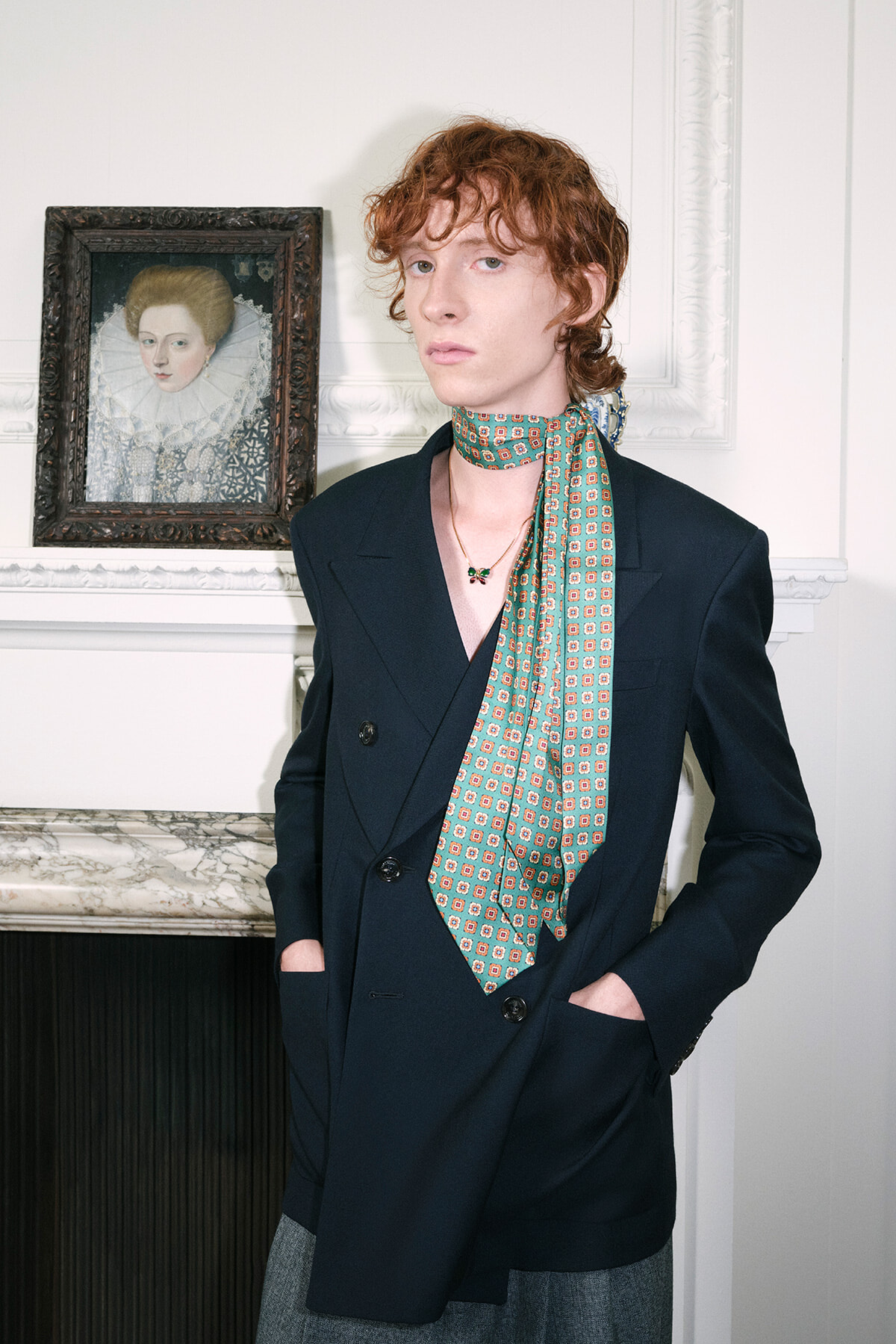 Young person with curly red hair in a navy blazer and teal patterned scarf, standing by a marble mantel with a framed portrait nearby.