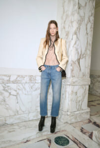Woman standing in a marble hallway wearing a beige blazer, pink lace top, blue jeans, black platform boots, layered necklace, and a chain bag.