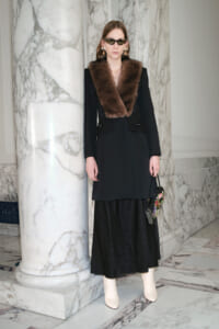 Model wearing a black tailored coat with a brown fur collar, black midi skirt, cream ankle boots, cat-eye sunglasses, and gold earrings, standing beside marble columns in a marble-filled interior.