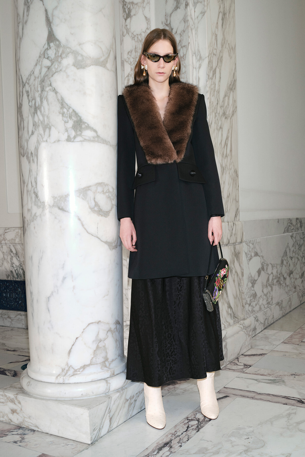 Model wearing a black tailored coat with a brown fur collar, black midi skirt, cream ankle boots, cat-eye sunglasses, and gold earrings, standing beside marble columns in a marble-filled interior.